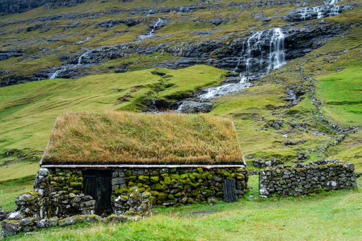 View of a traditional stone house with a turf roof nestled against a green hillside with a waterfall in the Faroe Islands.