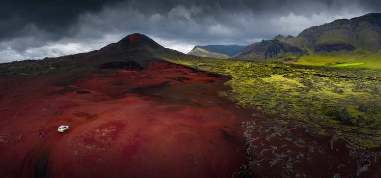 Aerial view of a white car driving through a dramatic volcanic landscape with red soil green mossy fields and rugged mountains under dark stormy clouds in Iceland.