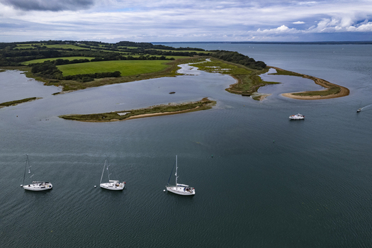 Aerial view of Newtown Creek with sailboats anchored in the calm water surrounded by lush green coastal landscape and tidal marshes in Isle of Wight United Kingdom.