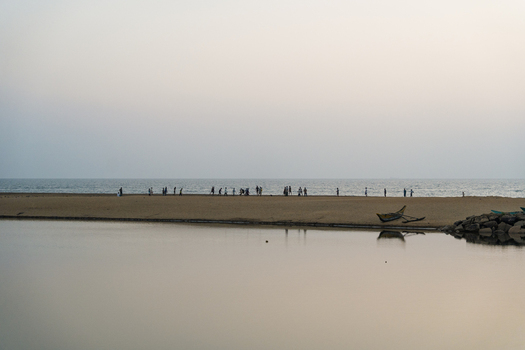 View of people walking along a narrow sandbar between a calm lagoon and the ocean with a small boat at dusk in Galle Southern Province Sri Lanka.