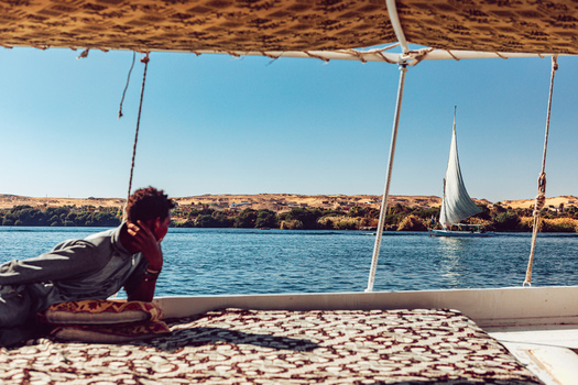 View of a person relaxing on a traditional boat looking out over the Nile river with a white sailboat in the distance near Aswan Aswan Governorate Egypt. View of a person relaxing on a traditional boat looking out over the Nile river with a white sailboat in the distance near Aswan Aswan Governorate Egypt.