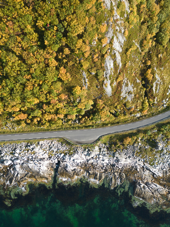 Aerial view of a coastal road winding between autumn colored forests and a rocky shoreline with turquoise water in Leknes Nordland Norway. Aerial view of a coastal road winding between autumn colored forests and a rocky shoreline with turquoise water in Leknes Nordland Norway.