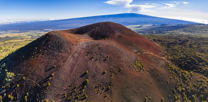 Aerial view of the Mauna Kea volcanic landscape featuring a prominent red cinder cone with sparse vegetation under a clear blue sky in Hawaii County Hawaii United States.