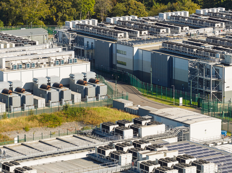 Aerial view of a large data center facility featuring multiple industrial buildings with rows of HVAC cooling units on the rooftops surrounded by security fencing and greenery in the United Kingdom.