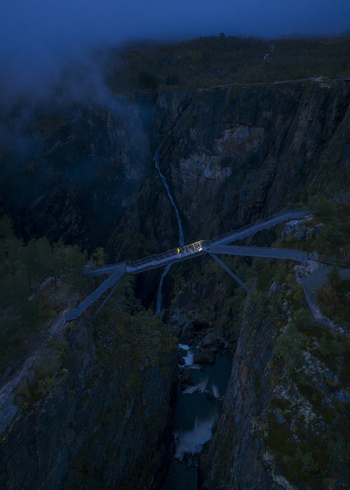 Aerial view of Voringfossen waterfall and the modern pedestrian bridge spanning the deep canyon with a person standing under the lights at dusk in Eidfjord Municipality Vestland Norway.