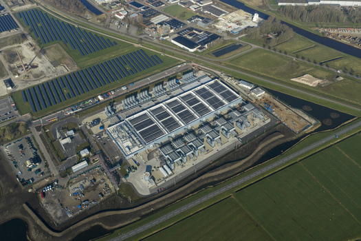 Aerial view of a digital data center facility with solar panels on the roof adjacent to a solar farm and green fields in Netherlands. Aerial view of a digital data center facility with solar panels on the roof adjacent to a solar farm and green fields in Netherlands.