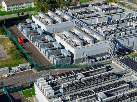 Aerial view of a large data center facility featuring rows of industrial cooling units on the rooftop surrounded by a secure fenced perimeter in the United Kingdom.