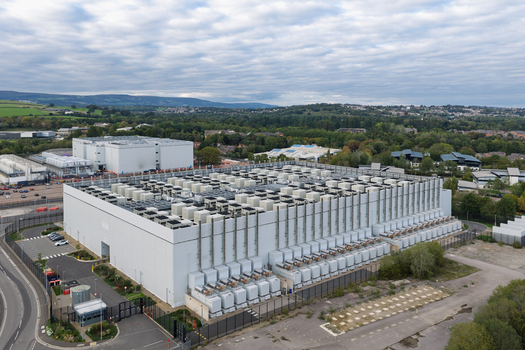 Aerial view of a large modern data center facility with numerous rooftop cooling units surrounded by a secure fence and green landscape in the United Kingdom.