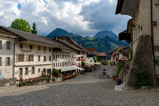 Gruyeres Switzerland 14 May 2026 View of the historic cobblestone main street lined with traditional buildings and restaurants against a backdrop of green mountains under a cloudy sky. Gruyeres Switzerland 14 May 2026 View of the historic cobblestone main street lined with traditional buildings and restaurants against a backdrop of green mountains under a cloudy sky.