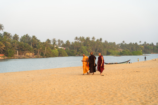 Sri Lanka 23 January 2026 View of three Buddhist monks walking along a sandy beach by a tropical lagoon with palm trees and a boat in the background.