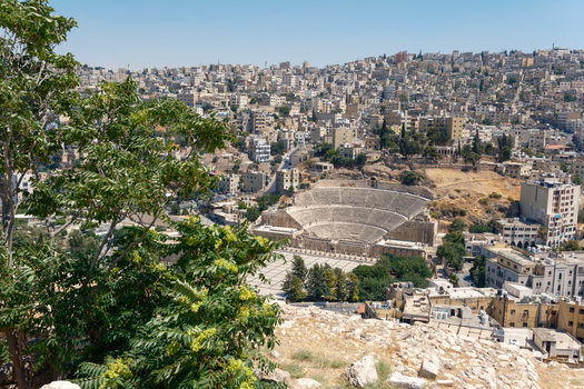 View of the historic Roman Theater nestled within the urban landscape featuring stone seating tiers and a large plaza in Amman Amman Governorate Jordan.