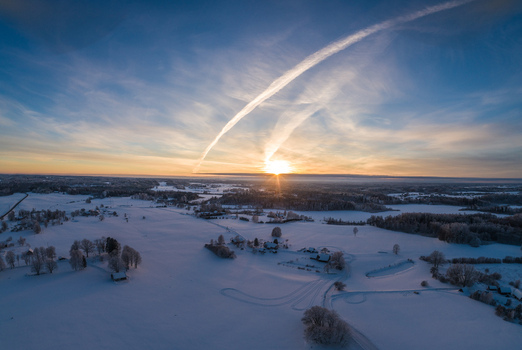 Aerial view of a snow covered landscape at sunset featuring frozen fields scattered trees and long contrails stretching across the sky in Latvia. Aerial view of a snow covered landscape at sunset featuring frozen fields scattered trees and long contrails stretching across the sky in Latvia.