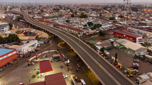 Aerial view of the urban landscape featuring a prominent overpass bridge bustling market areas with corrugated metal roofs and surrounding buildings at dusk in Tamale Northern Region Ghana. Aerial view of the urban landscape featuring a prominent overpass bridge bustling market areas with corrugated metal roofs and surrounding buildings at dusk in Tamale Northern Region Ghana.