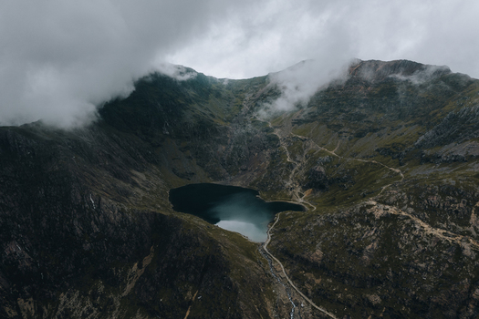 Aerial view of Snowdon and the dark waters of Llyn Glaslyn surrounded by rugged mountain slopes winding hiking trails and low hanging clouds in Caernarfon Wales United Kingdom.