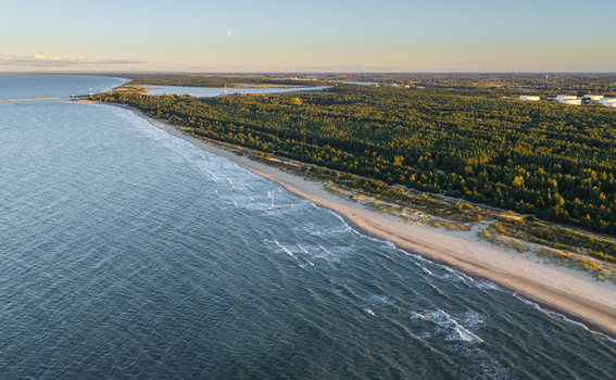 Aerial view of the Baltic Sea coastline featuring a sandy beach dense pine forest and industrial storage tanks in the distance in Gdansk Pomeranian Voivodeship Poland.