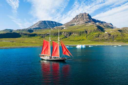 Aerial view of a sailboat with vibrant red sails navigating through deep blue Arctic waters surrounded by floating icebergs and a dramatic mountainous coastline in Greenland. Aerial view of a sailboat with vibrant red sails navigating through deep blue Arctic waters surrounded by floating icebergs and a dramatic mountainous coastline in Greenland.