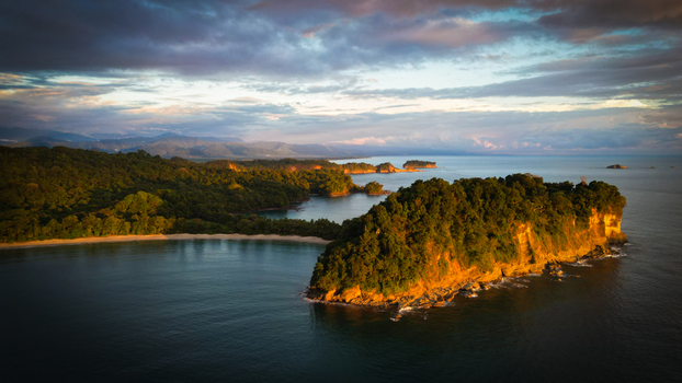 Aerial view of the rugged coastline kissed by golden sunlight meeting the tranquil dark sea Manuel Antonio National Park visible in the distance Quepos Puntarenas Province Costa Rica.