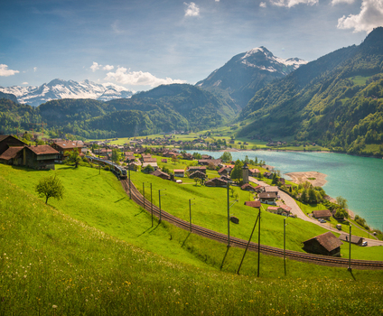 View of the Lungernersee lake with a train passing through the village of Lungern surrounded by lush green meadows and snow capped mountains in Lungern Obwalden Switzerland. View of the Lungernersee lake with a train passing through the village of Lungern surrounded by lush green meadows and snow capped mountains in Lungern Obwalden Switzerland.