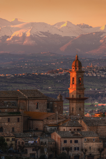 View of the historic bell tower and stone buildings of Morrovalle at sunset with snow capped mountains in the background Morrovalle Marche Italy.