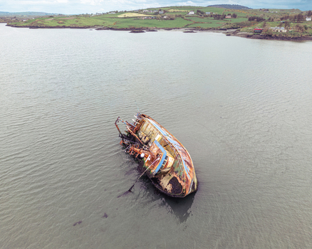 Aerial view of the abandoned MV Alta shipwreck resting in the shallow waters near the rocky coastline with its rusted hull and turquoise water against green fields Ireland. Aerial view of the abandoned MV Alta shipwreck resting in the shallow waters near the rocky coastline with its rusted hull and turquoise water against green fields Ireland.