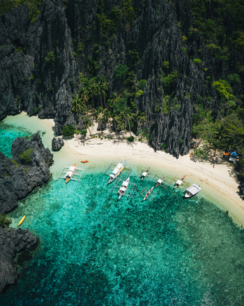 Aerial view of traditional outrigger boats anchored in turquoise waters near a white sand beach and towering limestone cliffs in El Nido Mimaropa Philippines. Aerial view of traditional outrigger boats anchored in turquoise waters near a white sand beach and towering limestone cliffs in El Nido Mimaropa Philippines.