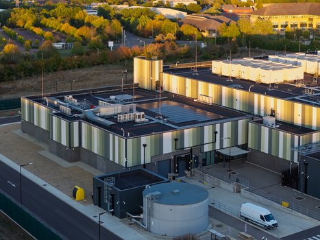 Aerial view of a modern data center facility with green and white striped panels rooftop HVAC units a large water tank and a white van parked in the yard United Kingdom. Aerial view of a modern data center facility with green and white striped panels rooftop HVAC units a large water tank and a white van parked in the yard United Kingdom.