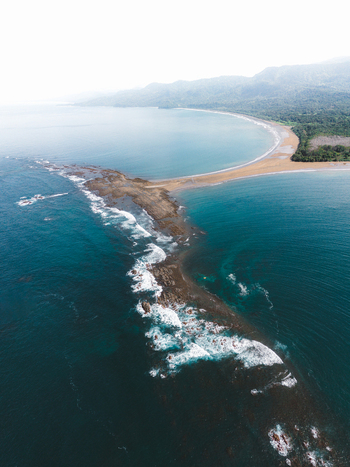 Aerial view of the Whale Tail rock formation and sandy beach at Marino Ballena National Park surrounded by turquoise ocean waves and lush tropical coastline in Costa Rica.