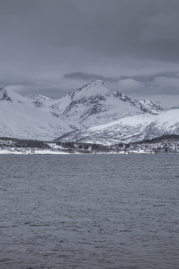 View of snow covered mountains rising above the cold waters of a fjord under a dark overcast sky Tromso Troms Norway.