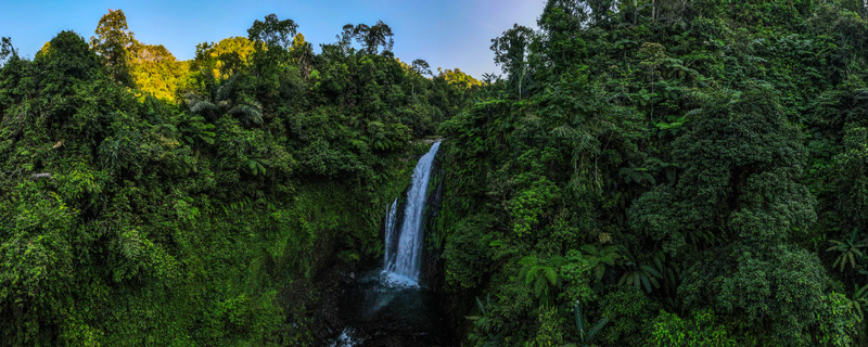 Aerial view of Gomblang Waterfall cascading down a rocky cliff surrounded by lush tropical jungle and dense green trees in Kecamatan Kedungbanteng Jawa Tengah Indonesia. Aerial view of Gomblang Waterfall cascading down a rocky cliff surrounded by lush tropical jungle and dense green trees in Kecamatan Kedungbanteng Jawa Tengah Indonesia.