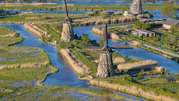 Aerial view of the historic Kinderdijk windmills standing along winding canals surrounded by lush green fields and reeds under bright sunlight Kinderdijk South Holland Netherlands. Aerial view of the historic Kinderdijk windmills standing along winding canals surrounded by lush green fields and reeds under bright sunlight Kinderdijk South Holland Netherlands.