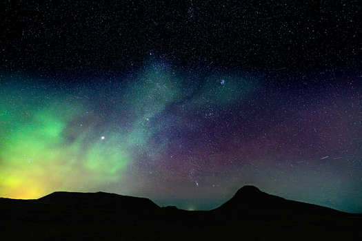 View of a surreal sky ablaze with ethereal green and purple hues dancing above the dark silhouettes of mountainous terrain Krysuvikurkirkja Iceland.
