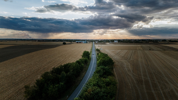 Aerial view of a straight road cutting through vast agricultural fields under a dramatic cloudy sky with sunbeams in Veliki Radinci Vojvodina Serbia.