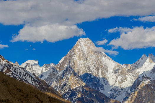 View of the snow capped peak of Gasherbrum IV under a bright blue sky with scattered clouds in Skardu Gilgit Pakistan. View of the snow capped peak of Gasherbrum IV under a bright blue sky with scattered clouds in Skardu Gilgit Pakistan.