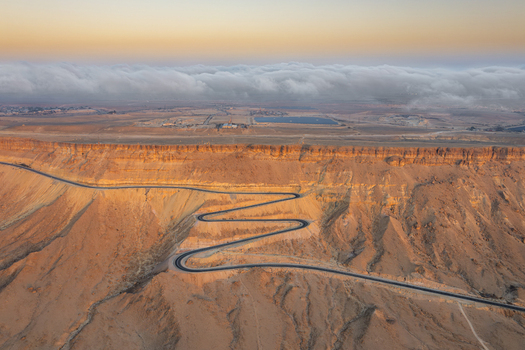 Aerial view of the winding road through the desert landscape of Makhtesh Ramon at sunset with low clouds over the crater rim in Mitzpe Ramon South District Israel. Aerial view of the winding road through the desert landscape of Makhtesh Ramon at sunset with low clouds over the crater rim in Mitzpe Ramon South District Israel.