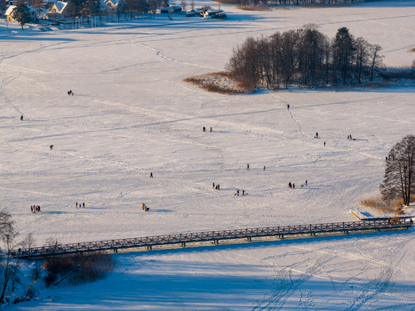 Aerial view of people walking on a frozen snow covered lake near a wooden bridge and small islands with bare trees in Lithuania. Aerial view of people walking on a frozen snow covered lake near a wooden bridge and small islands with bare trees in Lithuania.