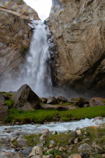 View of Khamosh Waterfall cascading down steep rocky cliffs into a flowing stream with green grass and boulders in Skardu Gilgit Pakistan.