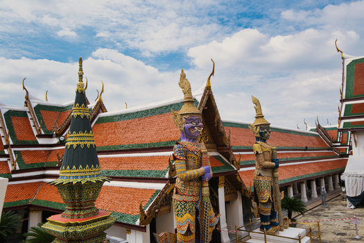 View of the ornate architecture and giant demon statues at The Grand Palace with traditional orange and green tiled roofs under a blue sky in Bangkok Thailand. View of the ornate architecture and giant demon statues at The Grand Palace with traditional orange and green tiled roofs under a blue sky in Bangkok Thailand.