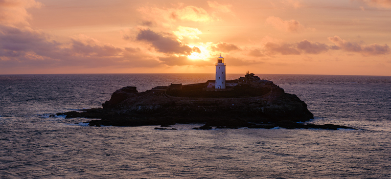 View of Godrevy Lighthouse on a rocky island during a fiery sunset with orange clouds over the Atlantic Ocean in Hayle Cornwall United Kingdom.