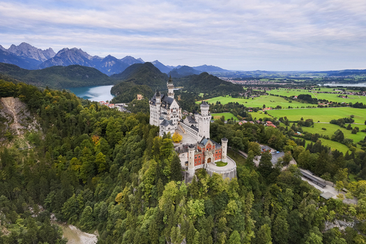 Aerial view of Neuschwanstein Castle perched majestically on a wooded hilltop overlooking a serene lake and the distant Alps Fussen Bayern Germany. Aerial view of Neuschwanstein Castle perched majestically on a wooded hilltop overlooking a serene lake and the distant Alps Fussen Bayern Germany.