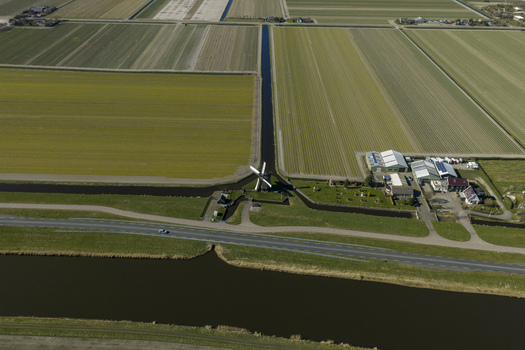 Aerial view of a traditional white windmill standing beside a straight canal amidst vast agricultural fields and farm buildings under bright daylight Netherlands. Aerial view of a traditional white windmill standing beside a straight canal amidst vast agricultural fields and farm buildings under bright daylight Netherlands.