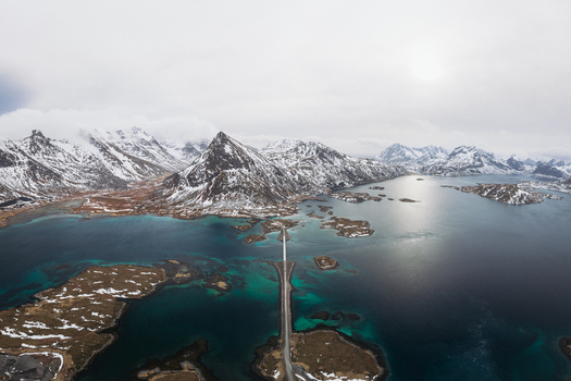 Aerial view of a long bridge stretching over turquoise waters connecting small islands beneath snow capped mountains Flakstadbruene Nordland Norway. Aerial view of a long bridge stretching over turquoise waters connecting small islands beneath snow capped mountains Flakstadbruene Nordland Norway.