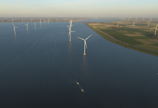Aerial view of an offshore wind farm with turbines standing in blue water next to green fields and a cargo barge in Netherlands. Aerial view of an offshore wind farm with turbines standing in blue water next to green fields and a cargo barge in Netherlands.
