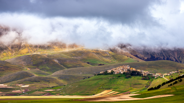 View of the village of Castelluccio di Norcia perched on a hill surrounded by the rolling green fields of the Piano Grande under a dramatic cloudy sky in Norcia Umbria Italy. View of the village of Castelluccio di Norcia perched on a hill surrounded by the rolling green fields of the Piano Grande under a dramatic cloudy sky in Norcia Umbria Italy.