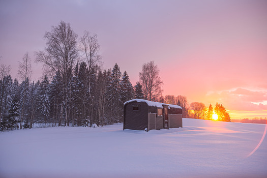 View of a small wooden cabin situated in a snow covered field surrounded by a dense forest of pine trees under a vibrant sunset sky in Latvia. View of a small wooden cabin situated in a snow covered field surrounded by a dense forest of pine trees under a vibrant sunset sky in Latvia.