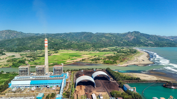 Aerial view of a power plant with a tall chimney and coal storage domes situated near the coast with green fields and mountains in the background Indonesia.