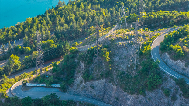 Aerial view of high voltage transmission towers and power lines crossing a winding mountain road near the turquoise waters of the lake in Rousset Serre Poncon Provence Alpes Cote d Azur France. Aerial view of high voltage transmission towers and power lines crossing a winding mountain road near the turquoise waters of the lake in Rousset Serre Poncon Provence Alpes Cote d Azur France.