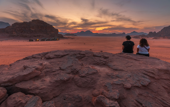 View of two people sitting on a rock overlooking the vast desert landscape of Wadi Rum Protected Area at sunset with distant tents in Wadi Rum Village Aqaba Governorate Jordan. View of two people sitting on a rock overlooking the vast desert landscape of Wadi Rum Protected Area at sunset with distant tents in Wadi Rum Village Aqaba Governorate Jordan.