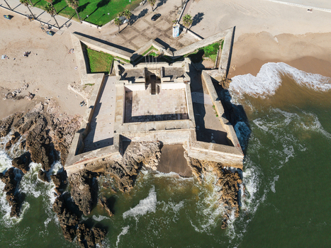 Aerial view of the star shaped Forte de Sao Bruno with waves crashing against its stone walls next to a sandy beach and palm trees in Caxias Lisbon Portugal.