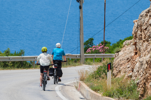 View of two cyclists with helmets and panniers riding on a winding coastal road overlooking the blue sea and rocky cliffs Vlore County Albania. View of two cyclists with helmets and panniers riding on a winding coastal road overlooking the blue sea and rocky cliffs Vlore County Albania.