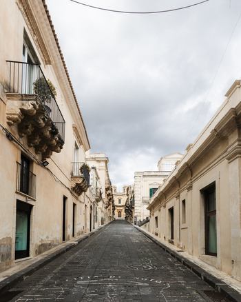 View of a historic narrow street with Baroque architecture and ornate balconies leading towards a church under an overcast sky in Noto Sicily Italy. View of a historic narrow street with Baroque architecture and ornate balconies leading towards a church under an overcast sky in Noto Sicily Italy.